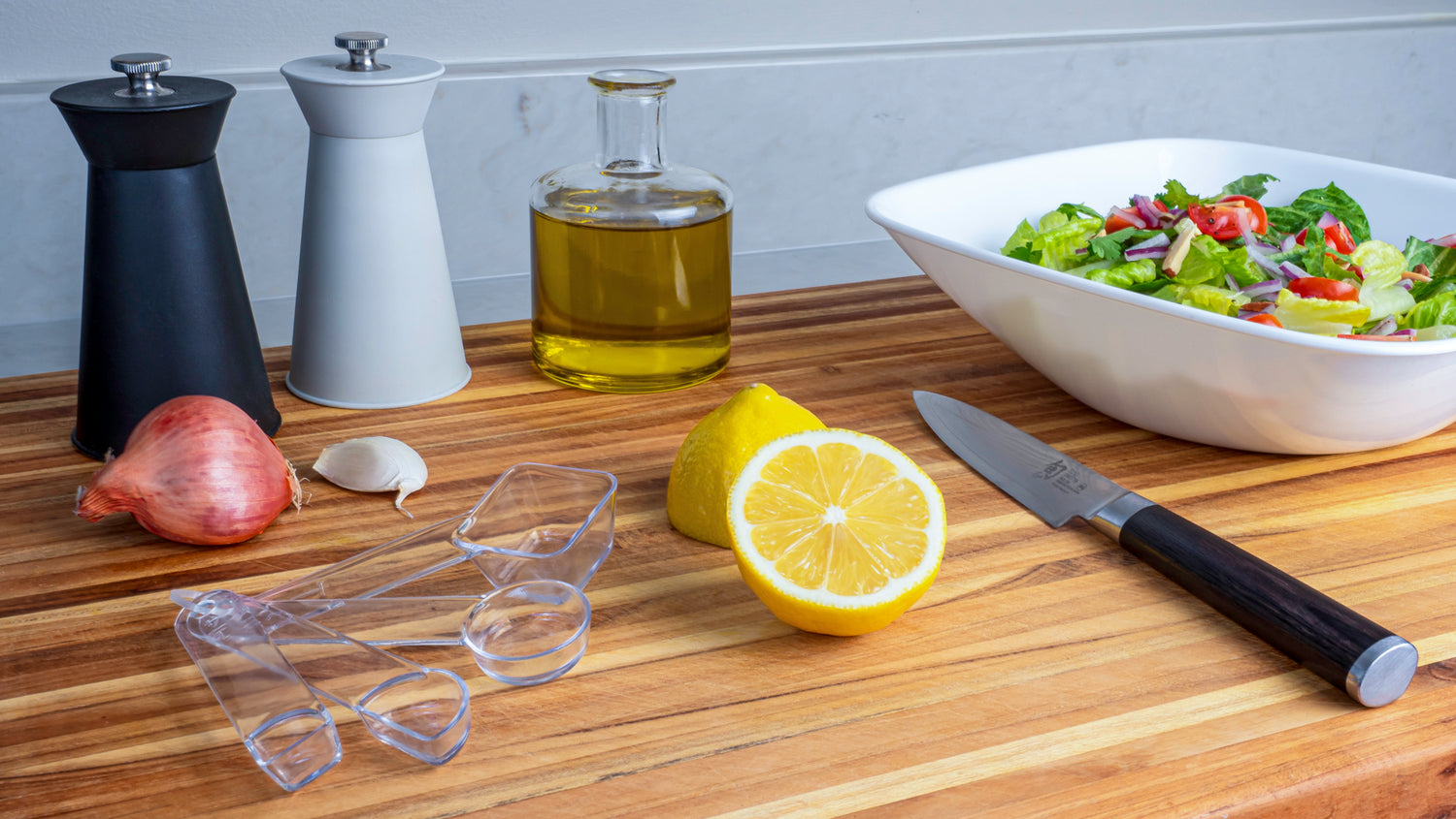 Photo of visual measuring spoons on a wooden cutting board with a lemon, knife, salad, seasonings and olive oil.